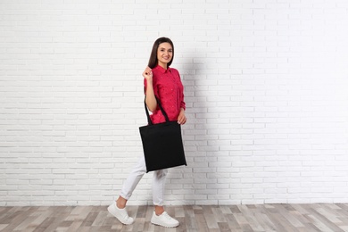 Photo of Beautiful young woman with stylish blank eco bag near brick wall, space for text