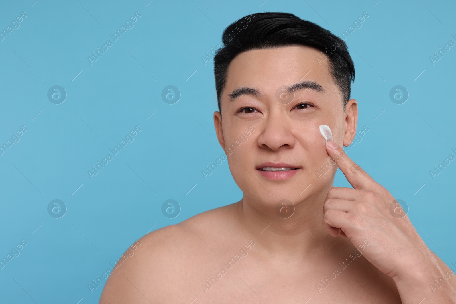 Photo of Handsome man applying cream onto his face on light blue background. Space for text