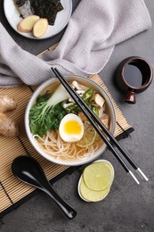 Photo of Bowl of vegetarian ramen, soy sauce, spoon and chopsticks on grey table, flat lay