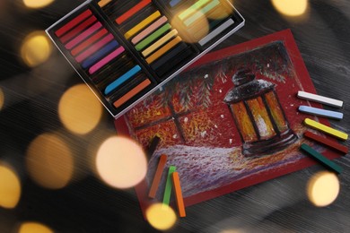 Photo of Colorful chalk pastels and beautiful painting on black wooden table, above view. Bokeh effect