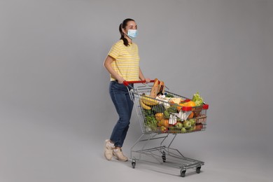 Photo of Woman with protective mask and shopping cart full of groceries on light grey background