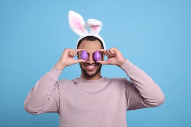 Photo of Happy African American man in bunny ears headband covering eyes with Easter eggs on light blue background