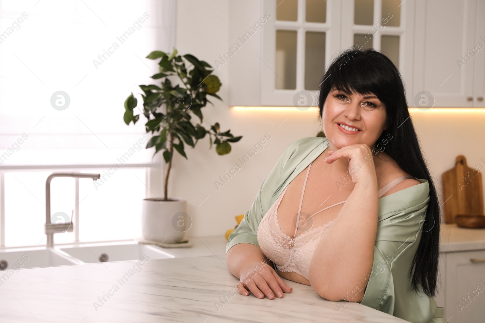 Photo of Beautiful overweight woman in beige underwear and silk robe at kitchen table. Plus-size model