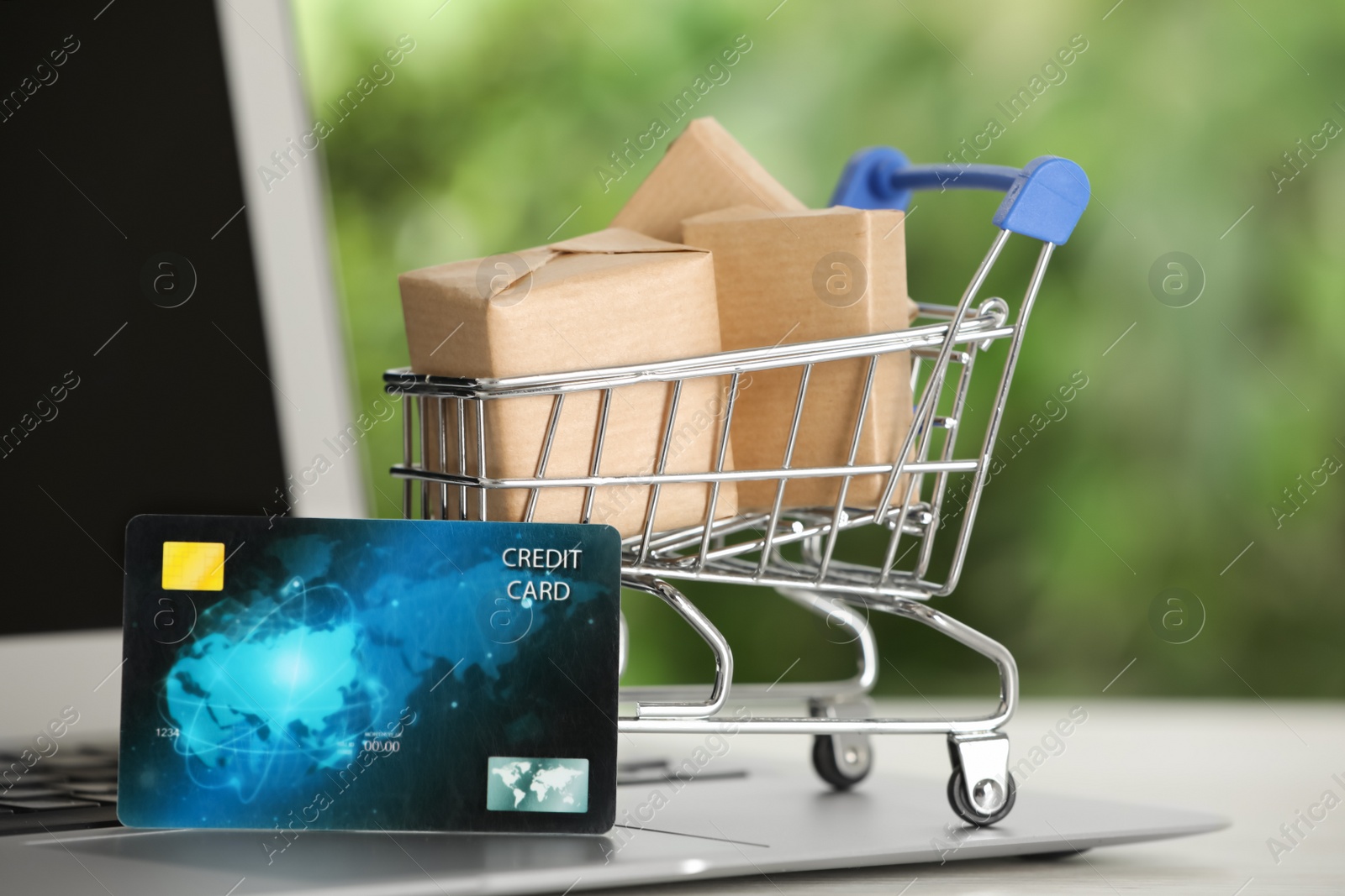 Photo of Online payment concept. Small shopping cart with bank card, boxes and laptop on table, closeup