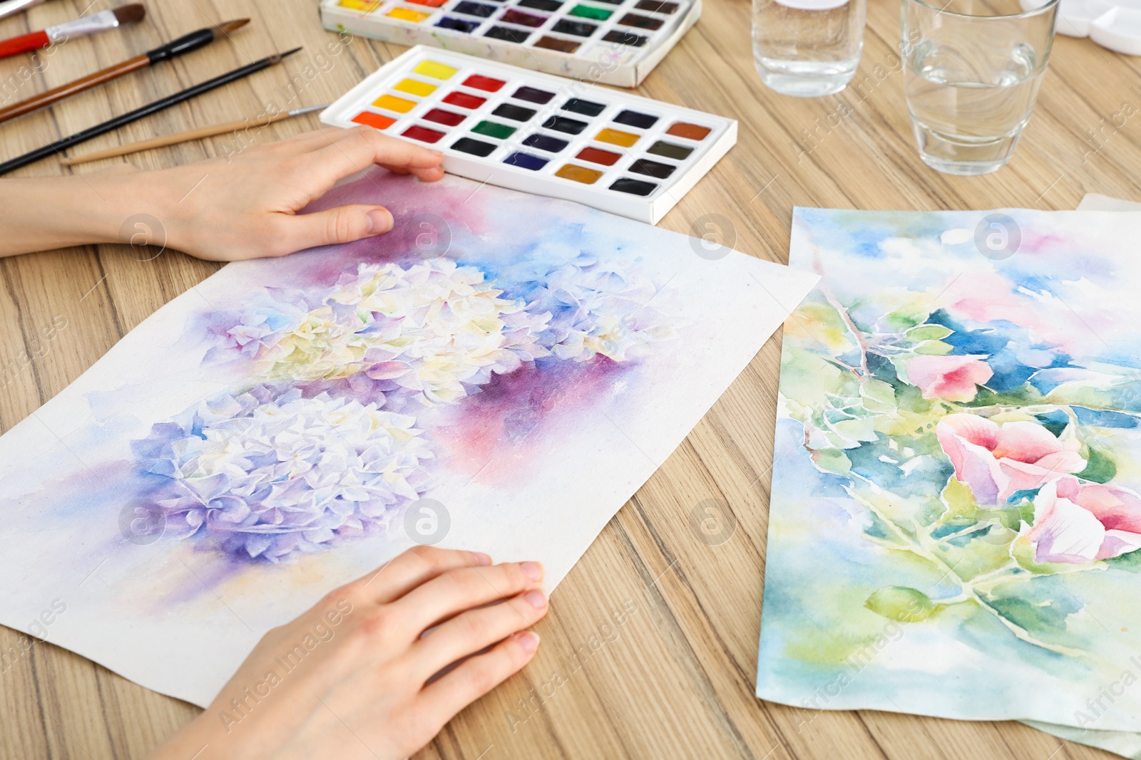 Photo of Woman holding painting of flowers near watercolor at wooden table, closeup. Creative artwork