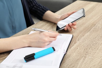 Photo of Woman taking notes while using smartphone at wooden table, closeup