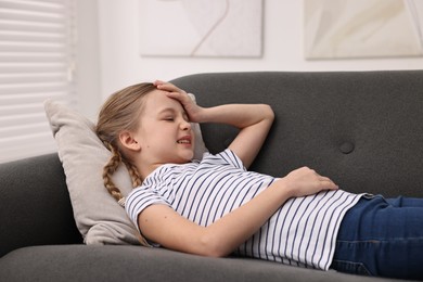 Photo of Little girl suffering from headache on sofa indoors