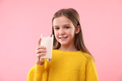 Happy little girl with milk mustache holding glass of tasty dairy drink on pink background