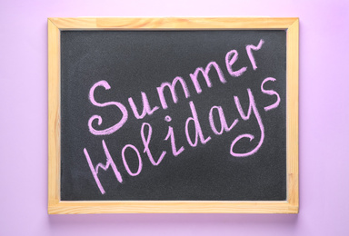 Chalkboard with phrase SUMMER HOLIDAYS on lilac background, top view. School's out