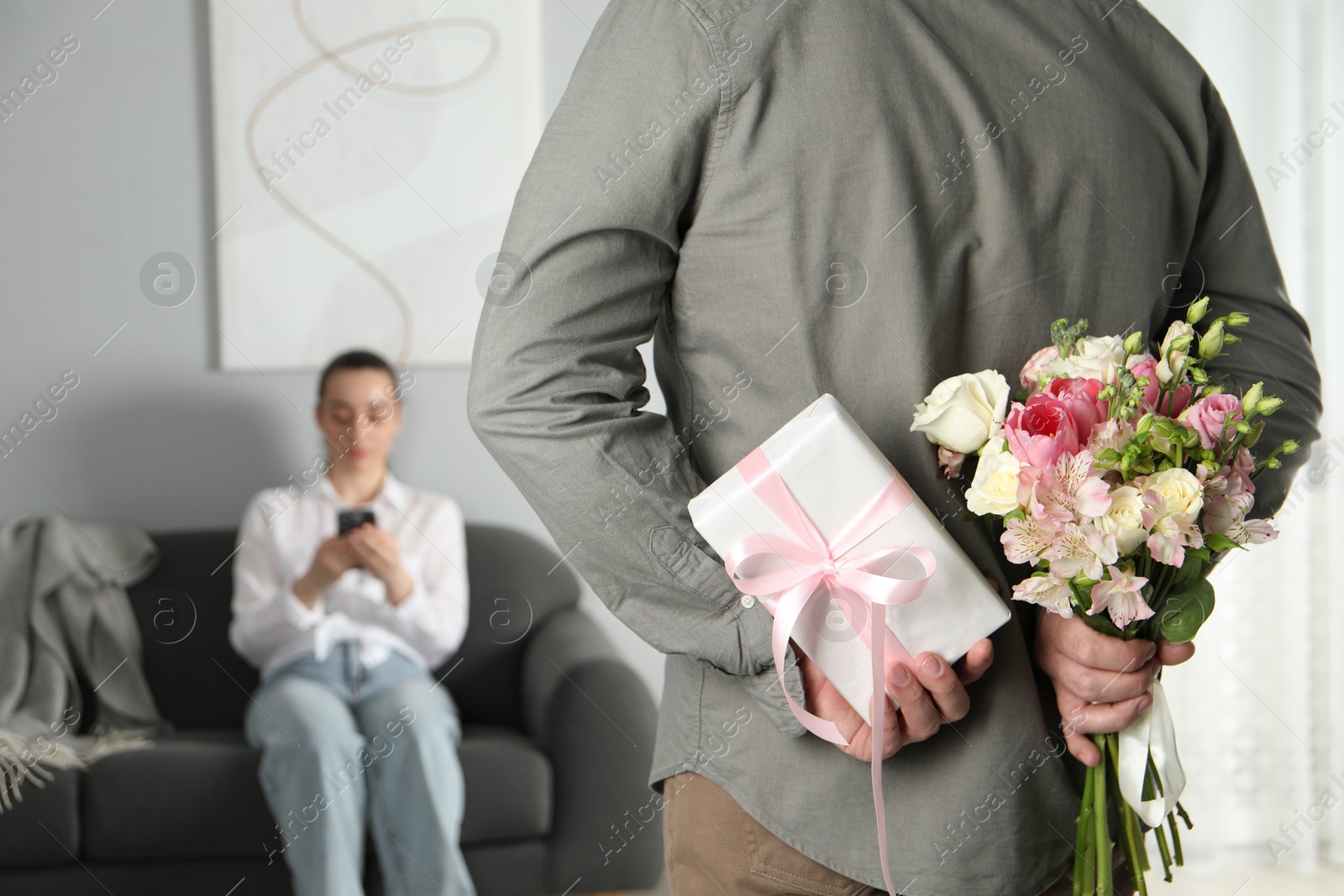 Photo of Man hiding bouquet of flowers and present for his beloved woman indoors, closeup