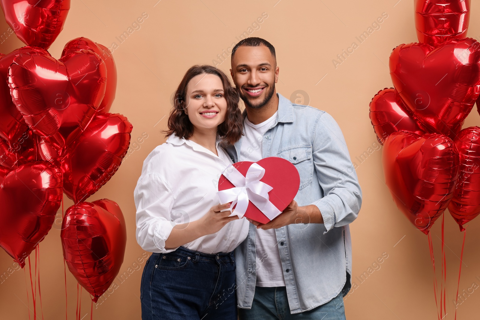 Photo of Lovely couple with gift box near heart shaped air balloons on beige background. Valentine's day celebration