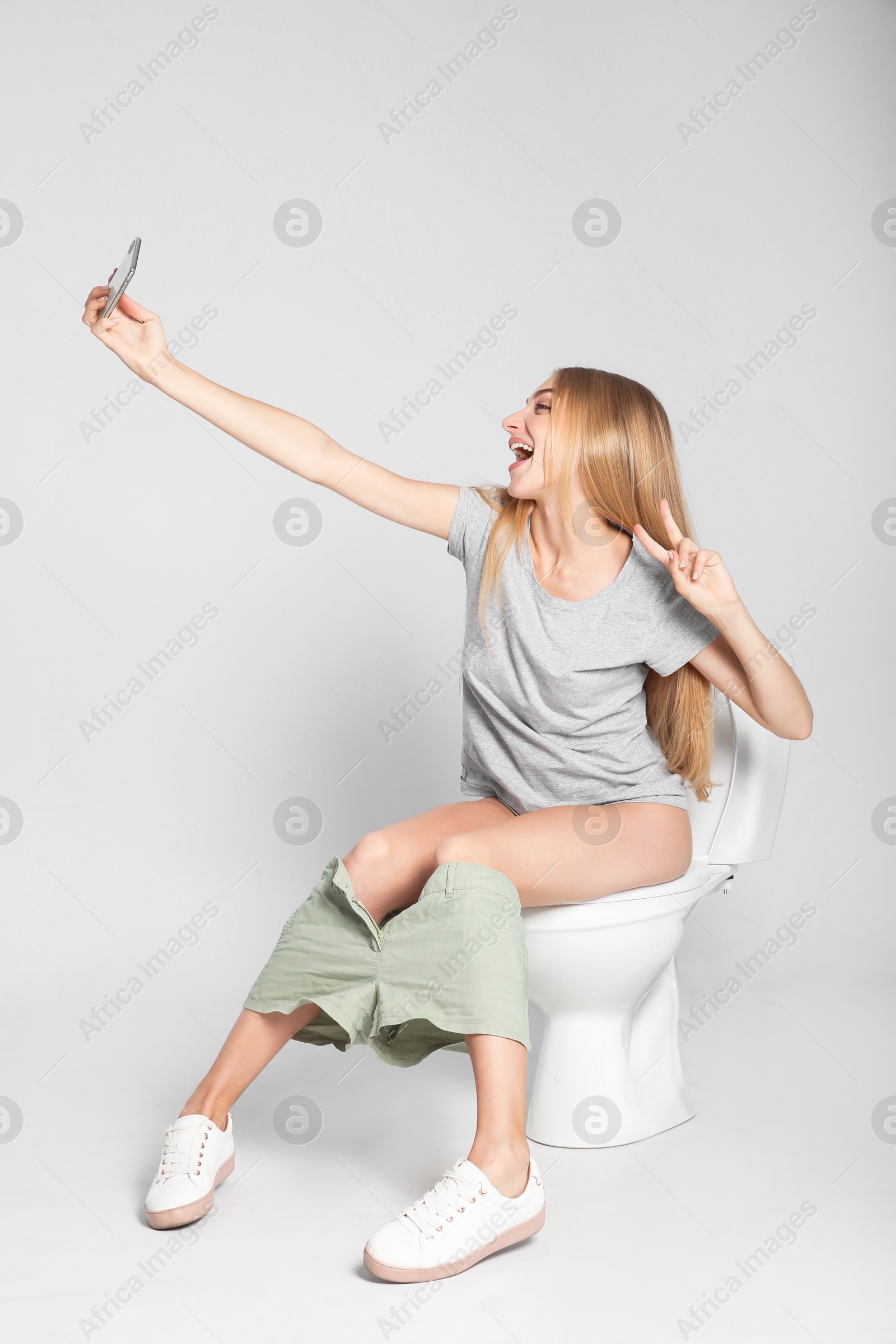 Photo of Young woman taking selfie while sitting on toilet bowl against gray background