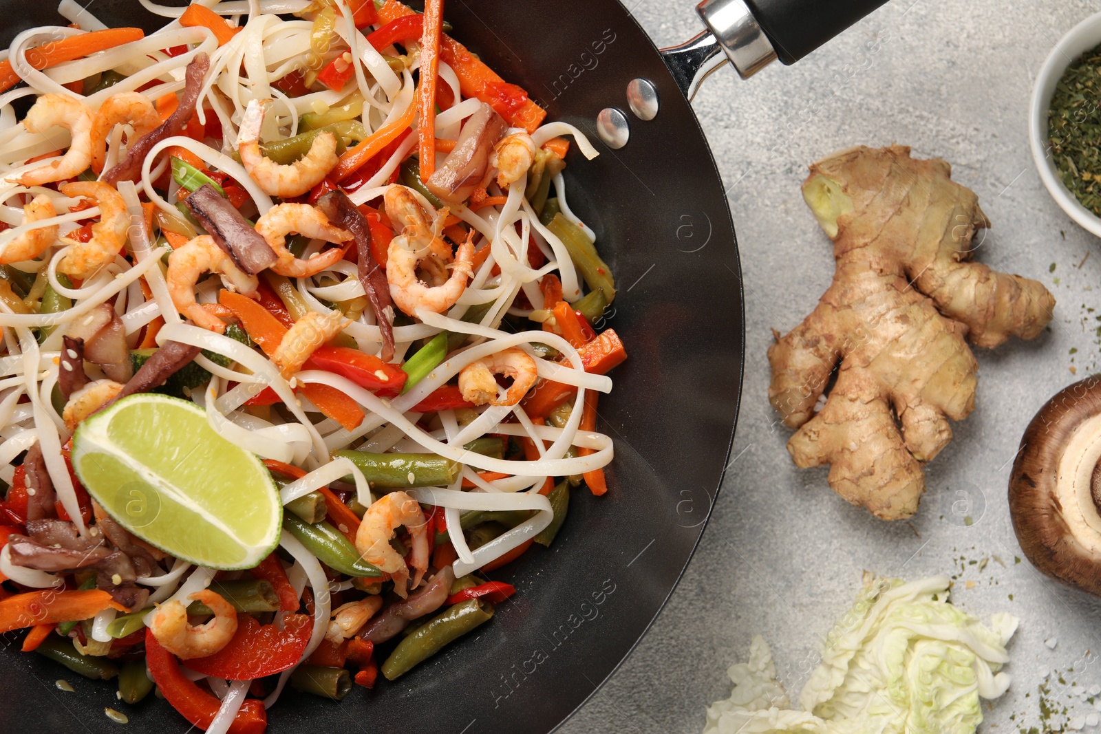 Photo of Shrimp stir fry with noodles and vegetables in wok surrounded by ingredients on grey table, flat lay