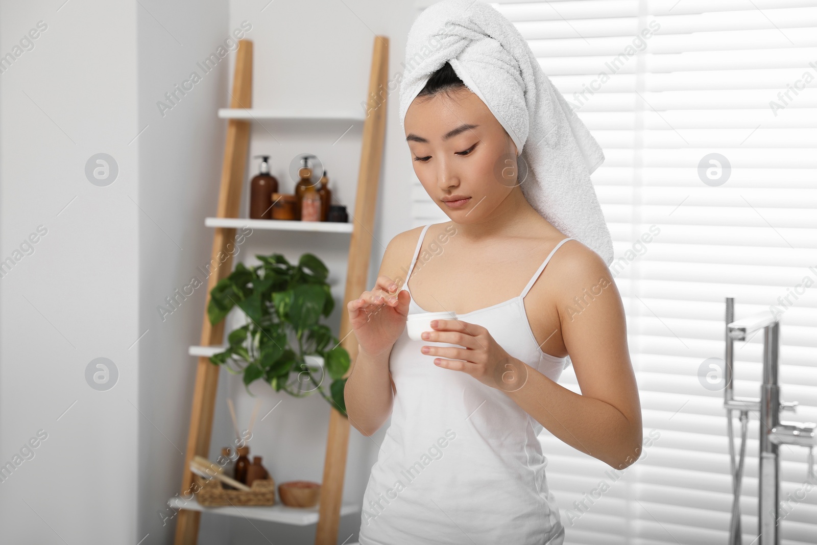 Photo of Beautiful young Asian woman holding body cream in room, space for text