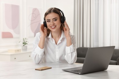 Photo of Happy woman with headphones and laptop at white table in room