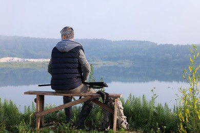 Man with hunting rifle sitting on wooden bench near lake outdoors, back view. Space for text