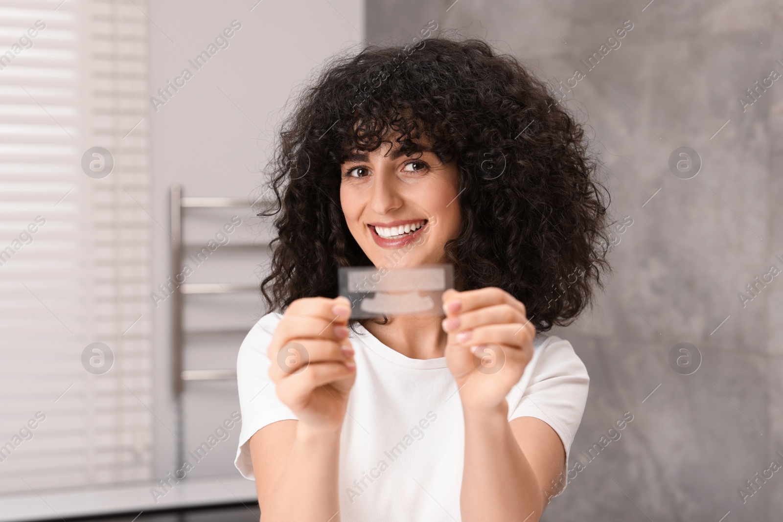 Photo of Young woman holding teeth whitening strips in bathroom