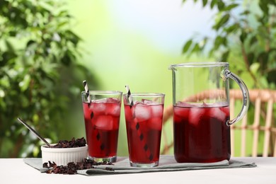 Photo of Refreshing hibiscus tea with ice cubes and dry roselle flowers on white table against blurred green background
