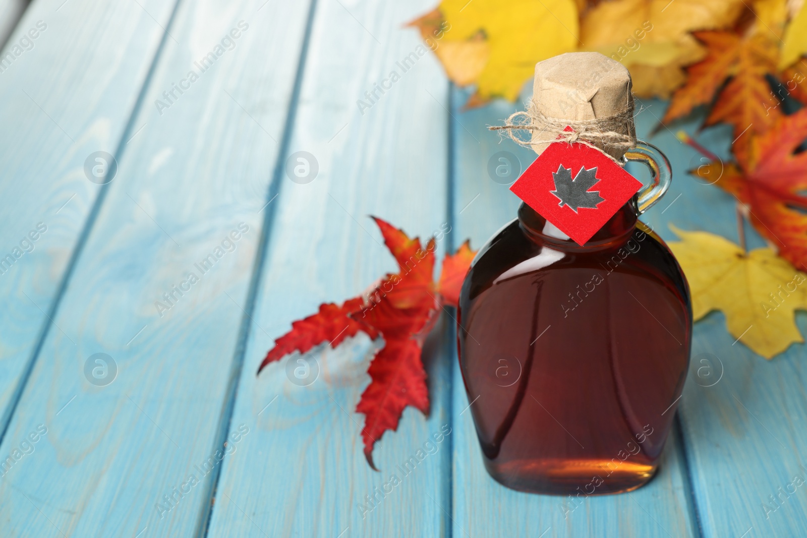 Photo of Glass bottle of tasty maple syrup and dry leaves on light blue wooden table. Space for text