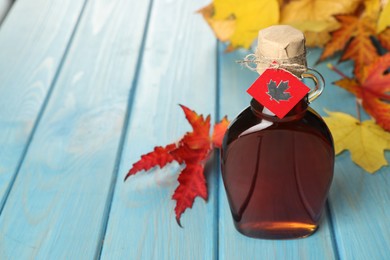 Photo of Glass bottle of tasty maple syrup and dry leaves on light blue wooden table. Space for text