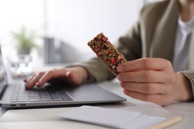 Photo of Woman holding tasty granola bar working with laptop at light table in office, closeup