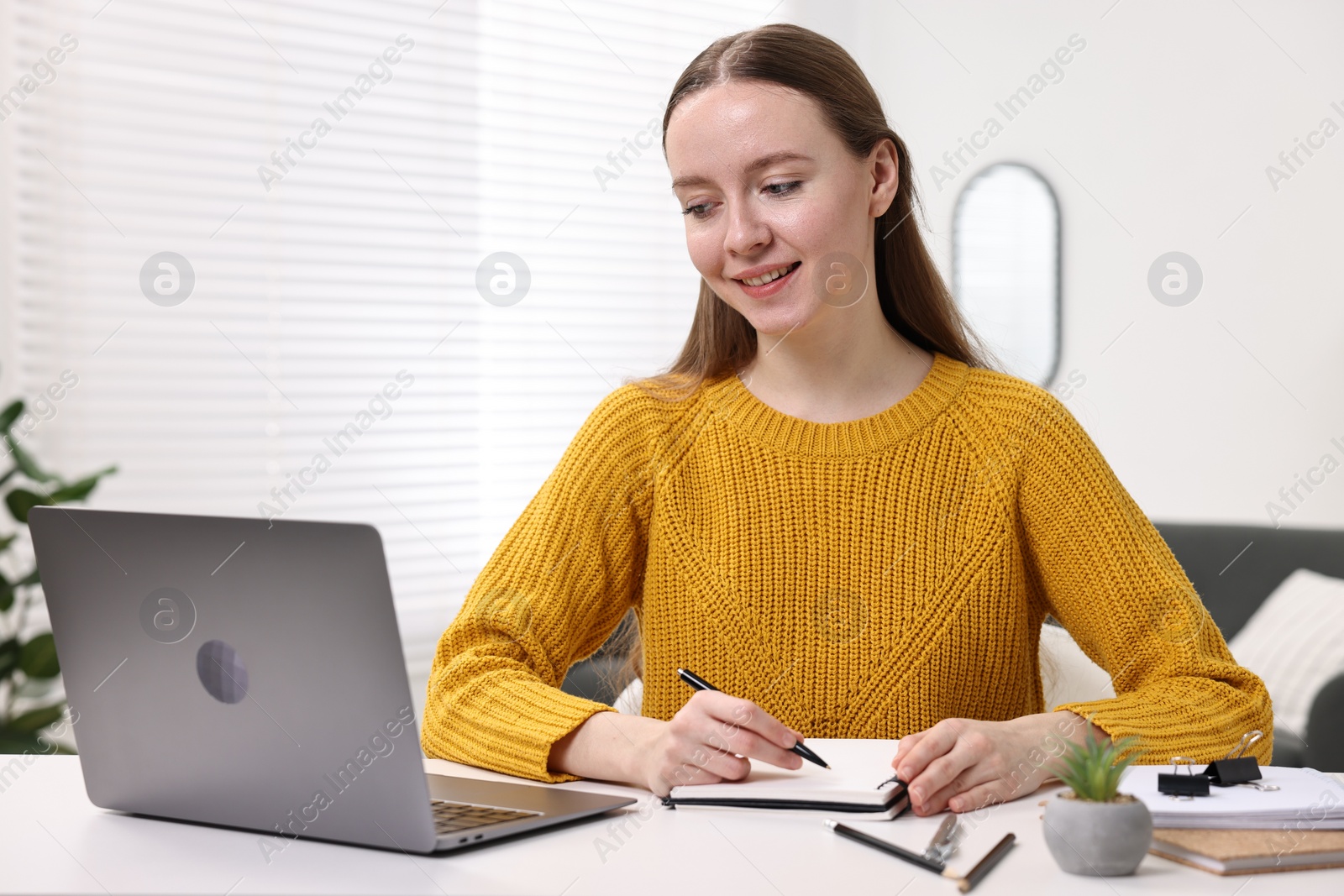 Photo of E-learning. Young woman taking notes during online lesson at white table indoors