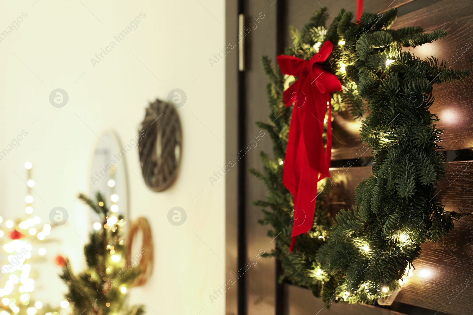 Photo of Beautiful Christmas wreath with red bow and festive lights hanging on door indoors. Space for text