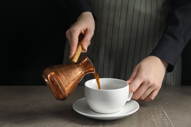 Photo of Turkish coffee. Woman pouring brewed beverage from cezve into cup at gray table against black background, closeup