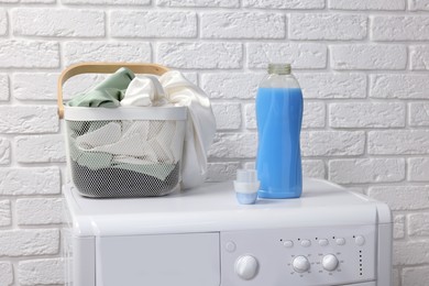 Bottle of fabric softener and basket with clothes on washing machine near white brick wall