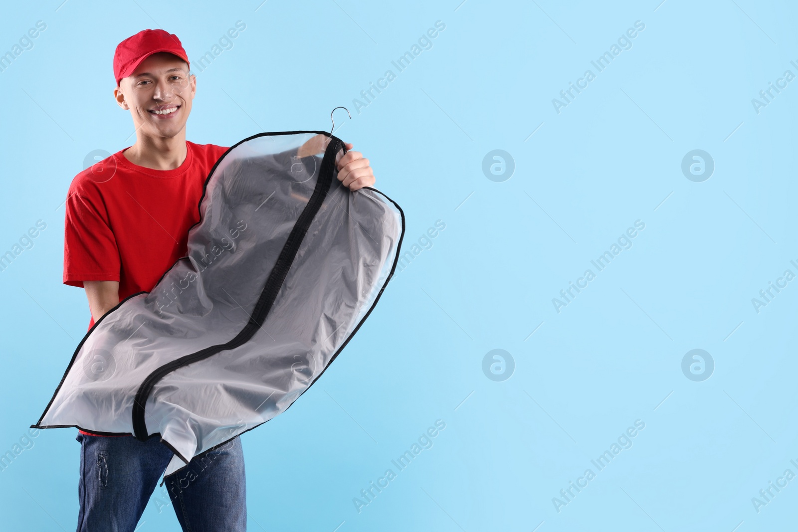 Photo of Dry-cleaning delivery. Happy courier holding garment cover with clothes on light blue background, space for text