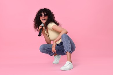 Beautiful young woman with microphone and sunglasses singing on pink background