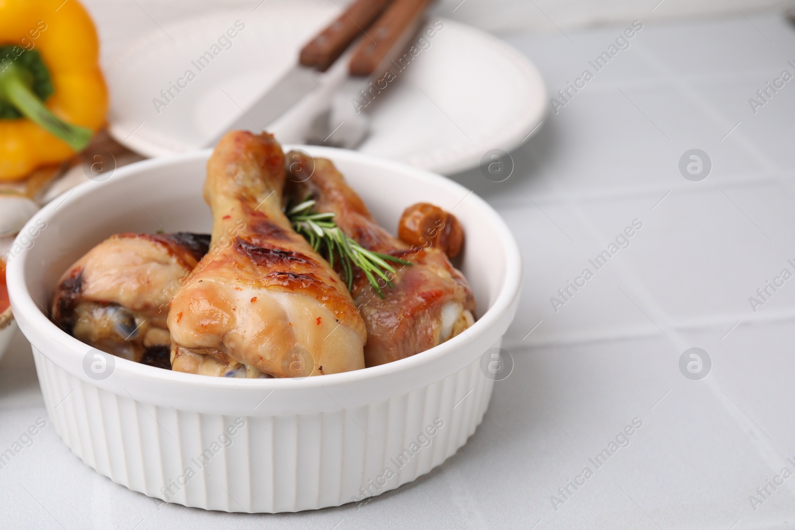 Photo of Delicious roasted chicken drumsticks with rosemary and tomatoes in bowl on white tiled table. Space for text