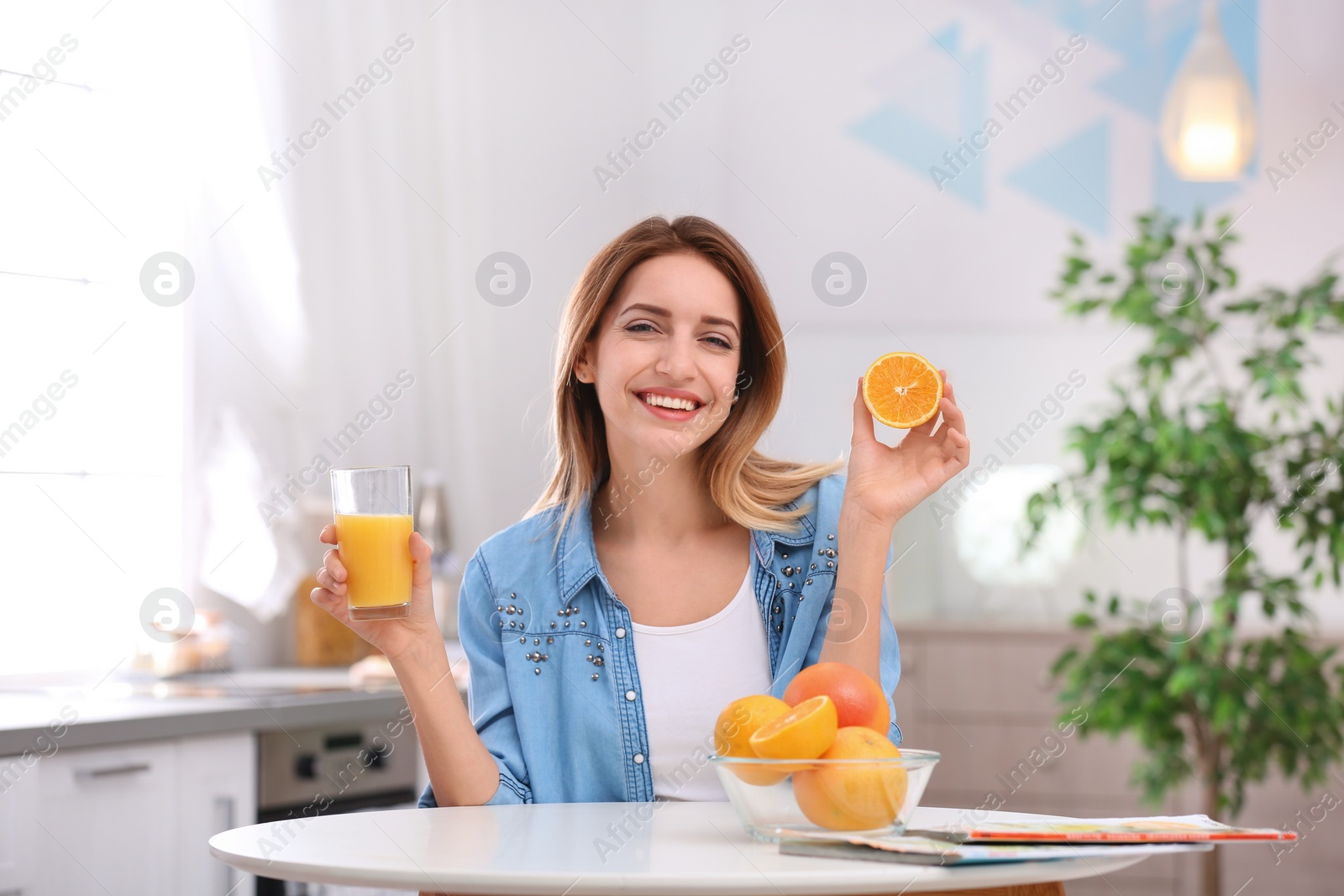 Photo of Happy young woman with glass of juice and orange at table in kitchen. Healthy diet