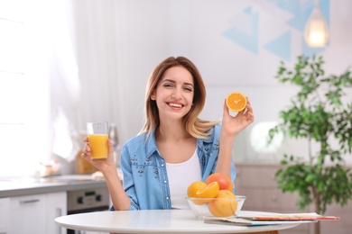 Photo of Happy young woman with glass of juice and orange at table in kitchen. Healthy diet