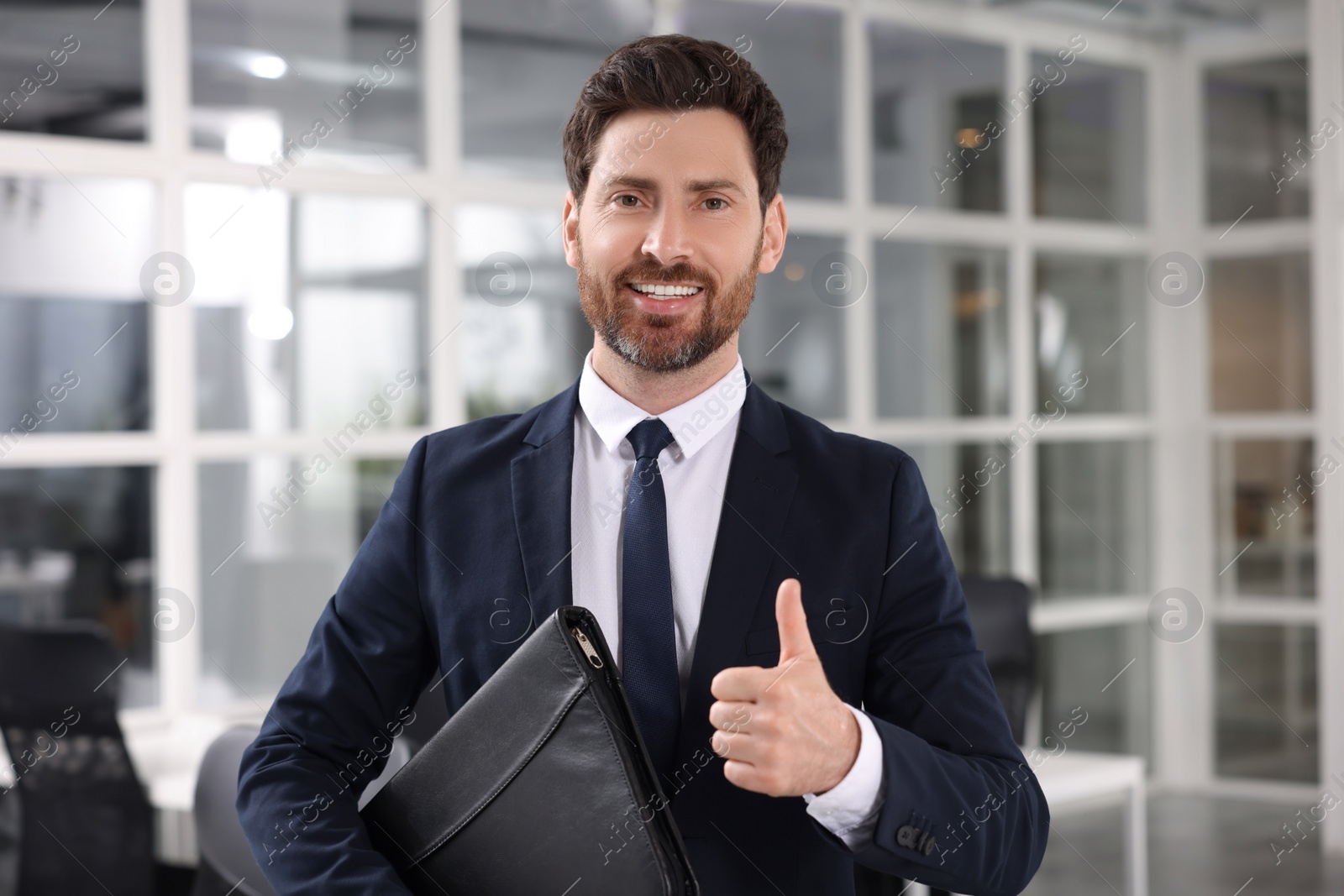 Photo of Happy real estate agent with leather portfolio showing thumb up indoors