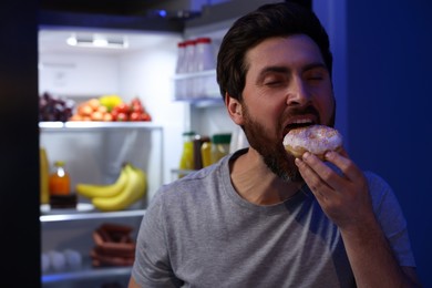 Man eating donut near refrigerator in kitchen at night, space for text. Bad habit