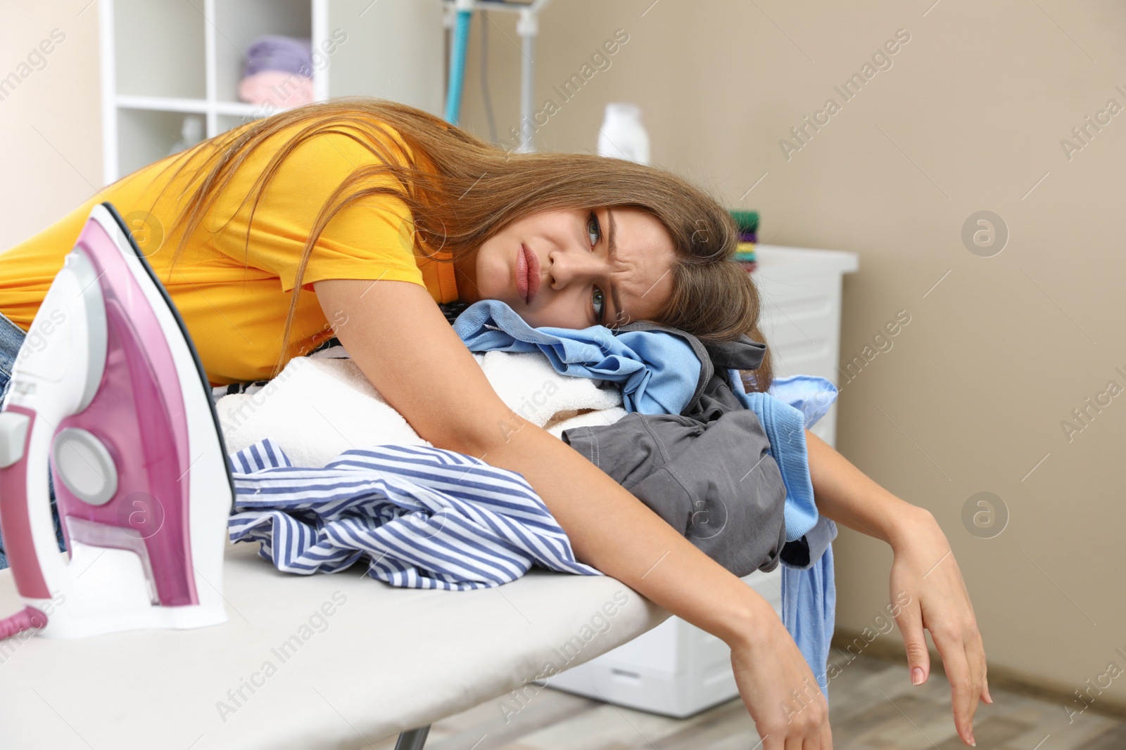 Photo of Tired woman leaning on ironing board with clothes at home