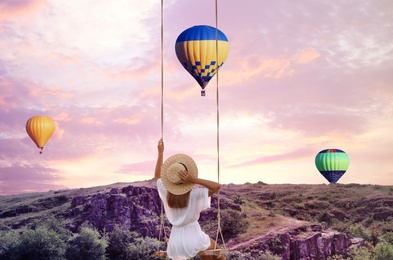 Dream world. Young woman swinging over mountains, hot air balloons in sunset sky on background