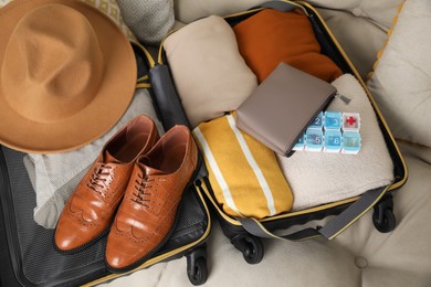 Open suitcase with packed clothes, accessories and pill box on sofa, above view