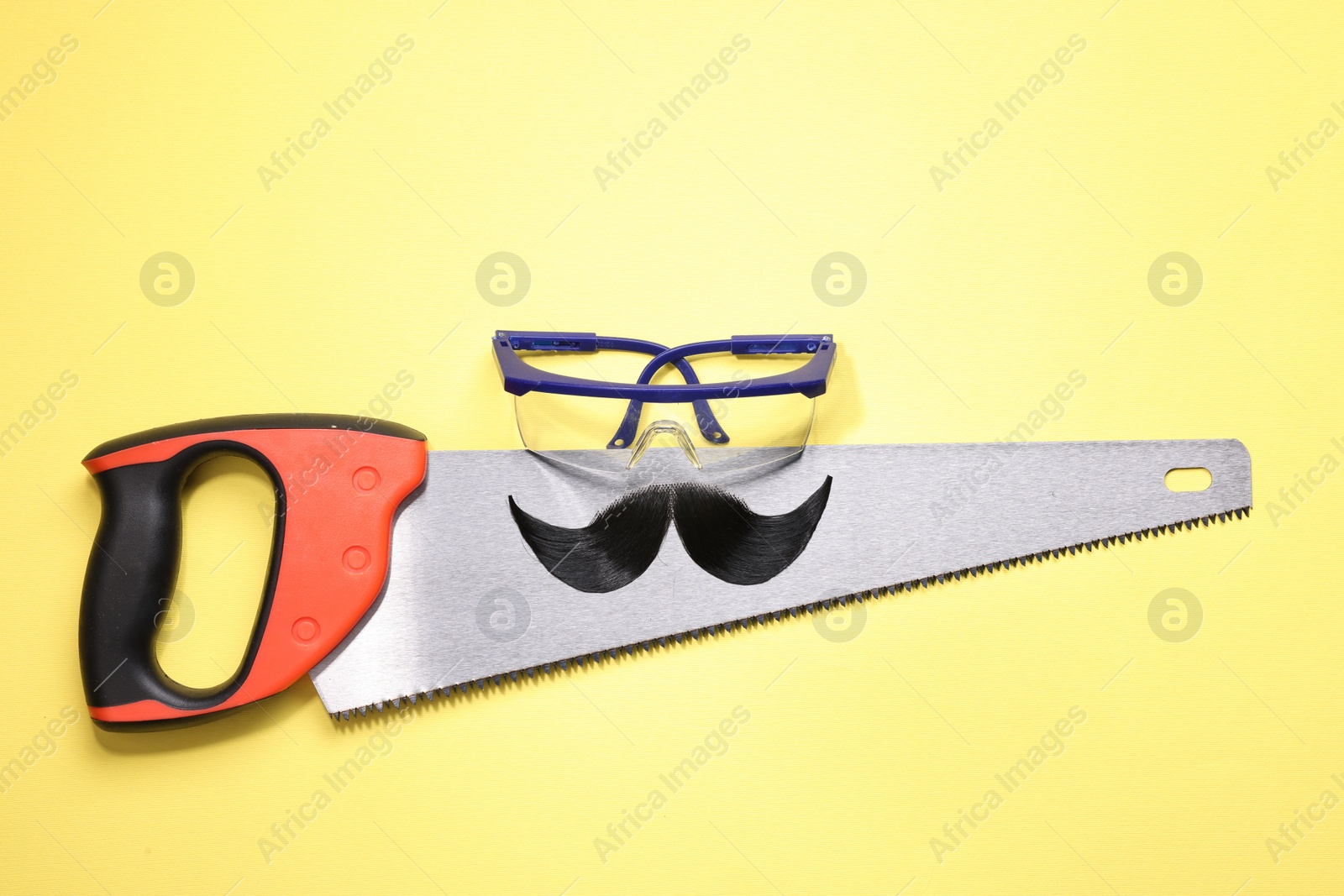 Photo of Man's face made of artificial mustache, safety glasses and hand saw on yellow background, top view