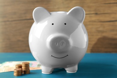 Ceramic piggy bank, banknotes and coins on blue wooden table, closeup