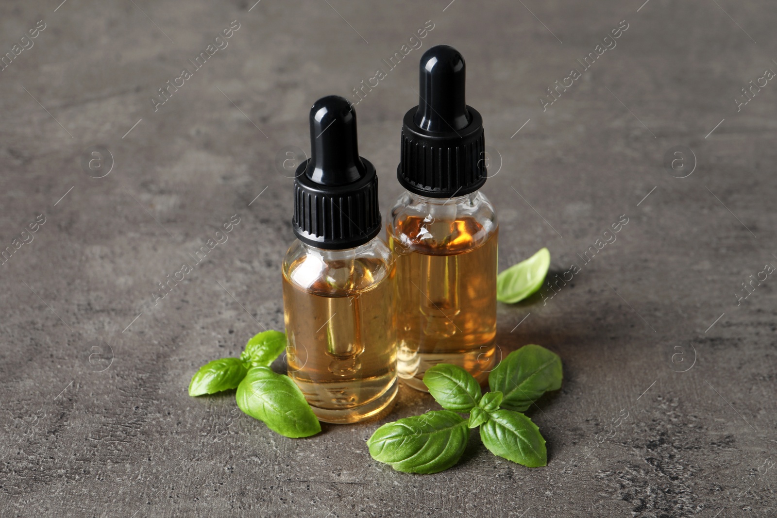 Photo of Glass bottles of basil essential oil and leaves on grey stone table