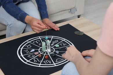 Photo of Astrologer predicting client's future with zodiac wheel, stones and crystals at wooden table indoors, closeup