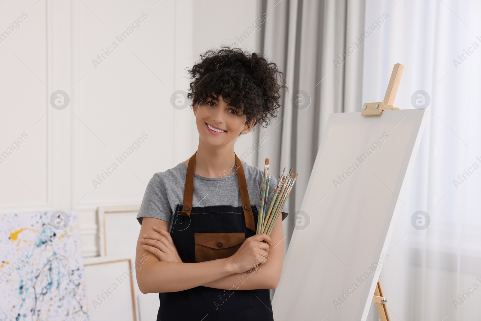 Photo of Young woman holding brushes near easel with canvas in studio