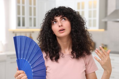 Photo of Young woman waving blue hand fan to cool herself in kitchen