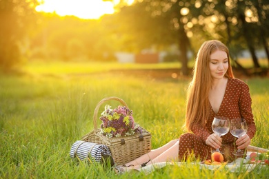 Photo of Beautiful young woman with wineglasses and picnic basket sitting on blanket in park