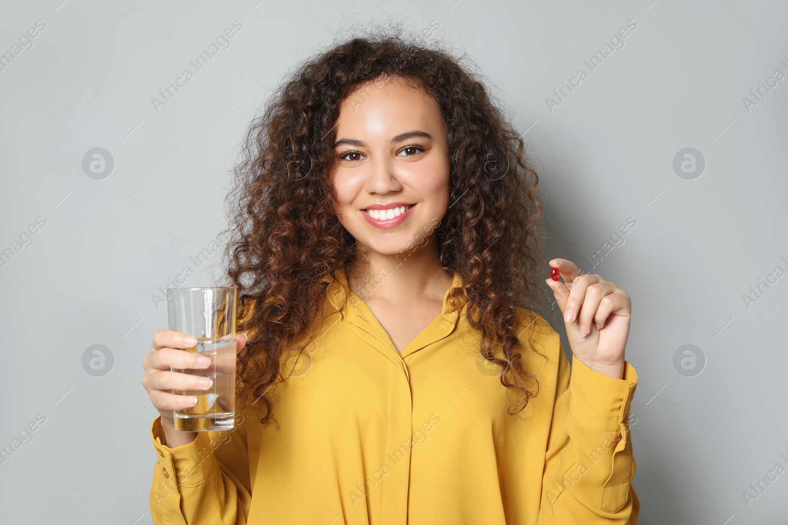 Photo of African-American woman with glass of water and vitamin capsule on light grey background