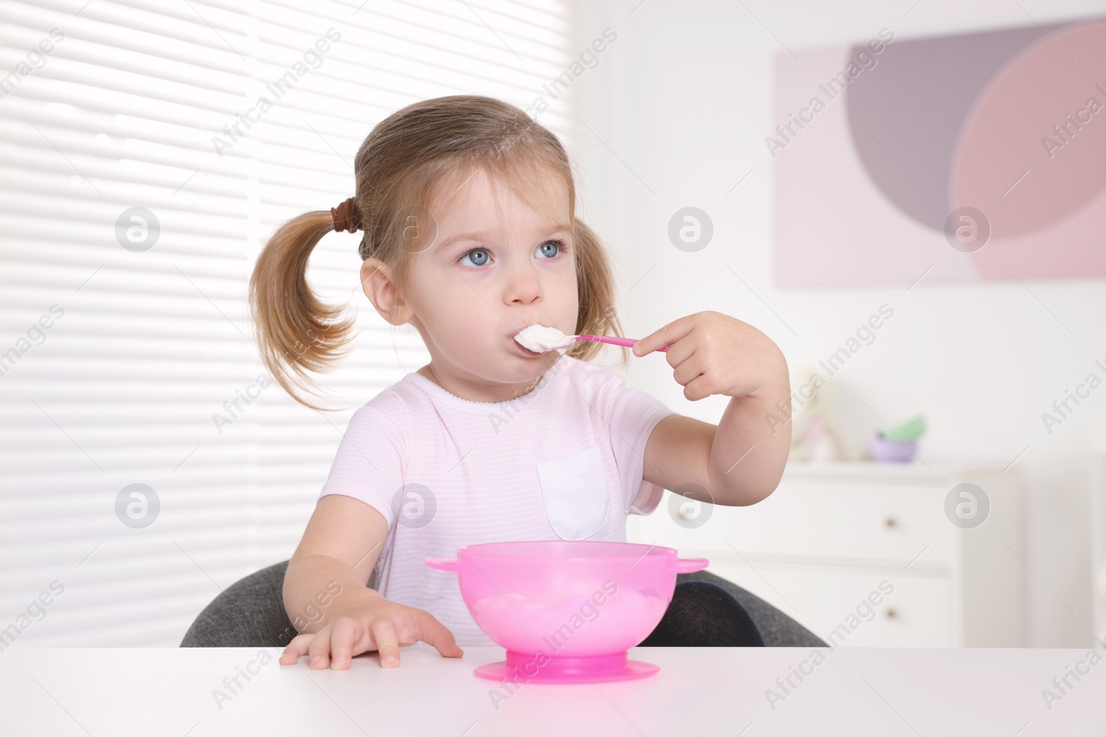 Photo of Cute little child eating tasty yogurt from plastic bowl with spoon at white table indoors