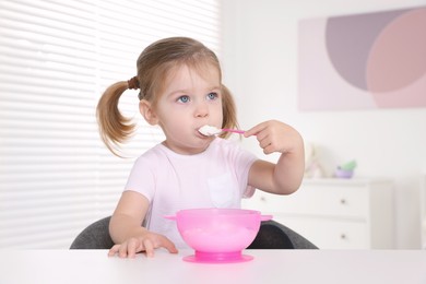 Cute little child eating tasty yogurt from plastic bowl with spoon at white table indoors