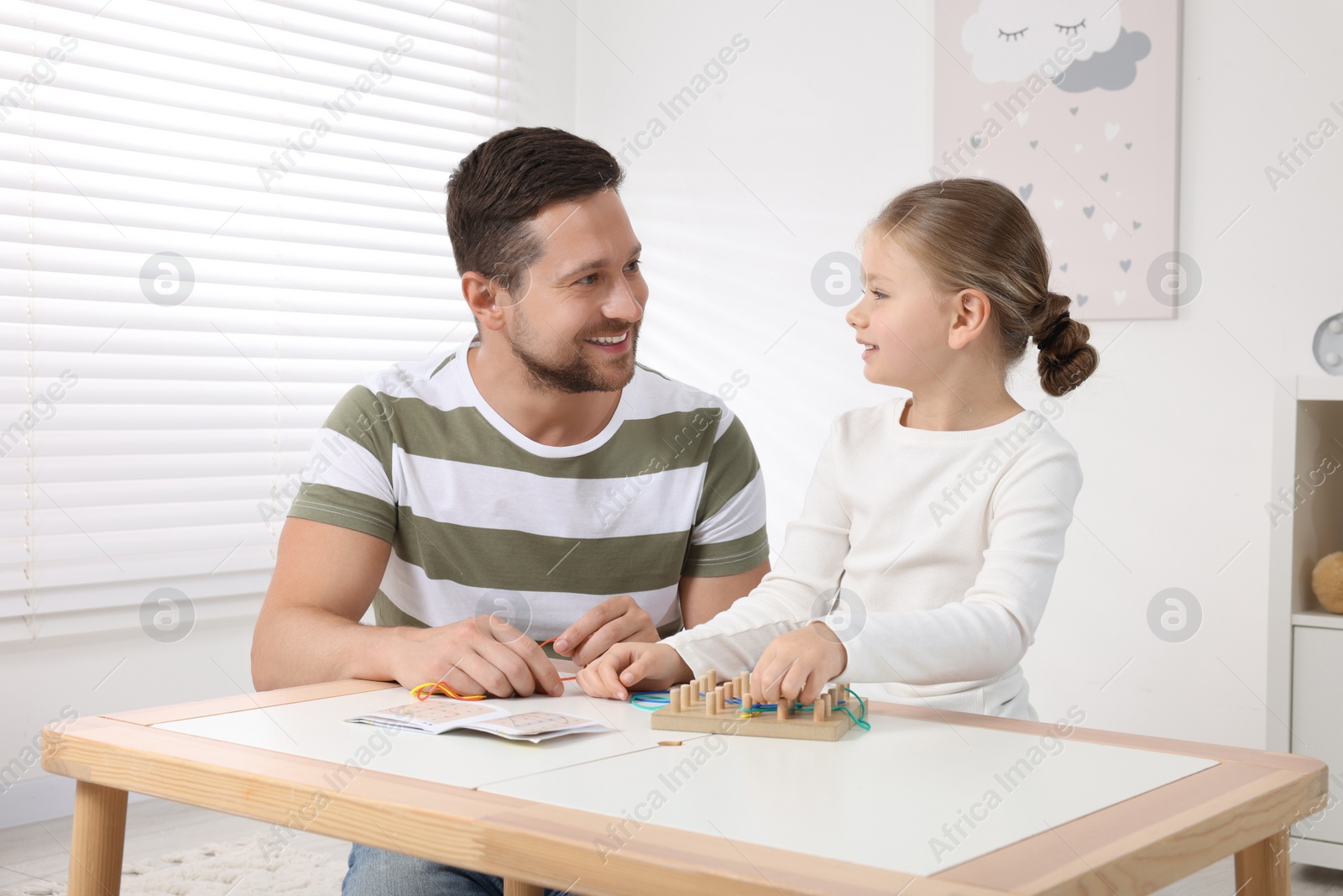 Photo of Motor skills development. Father helping his daughter to play with geoboard and rubber bands at white table in room